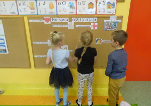 Troje dzieci stoi pod tablicą i przywiesza ilustracje pasujące do konkretnego prawa dziecka.